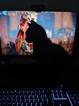 A cat is sitting in front of a computer screen, partially obscuring a video game character. The character has red hair and a white headdress. A backlit keyboard is visible at the bottom of the image, creating a gaming setup ambiance.