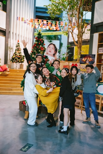 A group of eight people enjoying a festive gathering indoors. Several of them are dressed in casual clothing with holiday-themed accessories. They are standing in front of decorated Christmas trees, and one person is playfully holding up another. The room is adorned with colorful flags and holiday decorations, adding to the cheerful atmosphere.