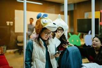 People are gathered in a room, some wearing unique animal hats and holding phones. The setting resembles an office space with glass walls, a whiteboard, and various tech-related decorations on the walls. The group appears to be engaged in a casual and playful activity.