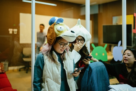 People are gathered in a room, some wearing unique animal hats and holding phones. The setting resembles an office space with glass walls, a whiteboard, and various tech-related decorations on the walls. The group appears to be engaged in a casual and playful activity.