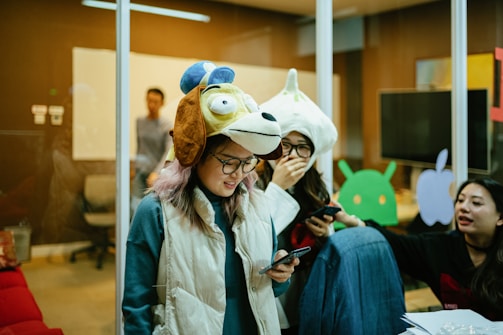 People are gathered in a room, some wearing unique animal hats and holding phones. The setting resembles an office space with glass walls, a whiteboard, and various tech-related decorations on the walls. The group appears to be engaged in a casual and playful activity.