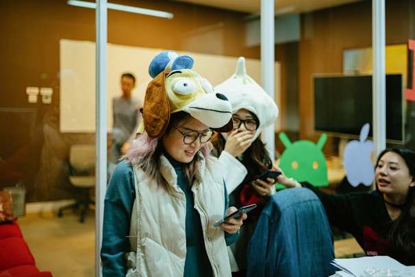 People are gathered in a room, some wearing unique animal hats and holding phones. The setting resembles an office space with glass walls, a whiteboard, and various tech-related decorations on the walls. The group appears to be engaged in a casual and playful activity.