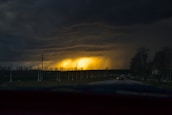 A dark stormy sky dominates the scene with a glowing, bright yellow light in the center, resembling a sunset or break in the storm clouds. The foreground features a road lined with power lines and sparse, leafless trees, contributing to a dramatic visual effect.