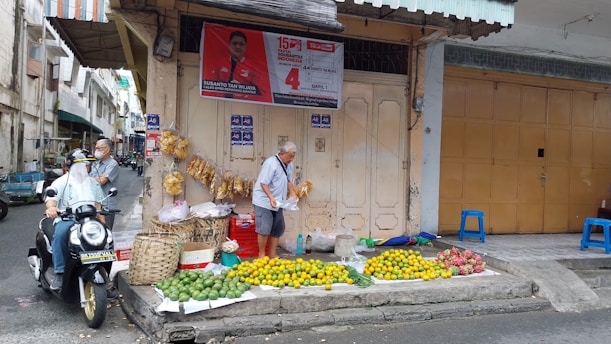A street scene with a small fruit stand displaying green and yellow fruits on the ground. There are baskets and bags attached to the wall beside the stand. A man is adjusting items on the stand, while another individual wearing a mask sits on a scooter nearby. A political campaign poster is visible above the stand, and the background features an urban environment with buildings and narrow streets.