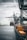 A large cargo ship is docked at an industrial port with towering cranes. Shipping containers are stacked on the ship and along the dock. The sky is overcast, creating a muted ambiance over the water.