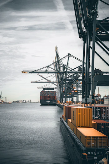 A large cargo ship loaded with containers docked at a busy port under overcast skies.