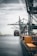 A large cargo ship is docked at an industrial port with towering cranes. Shipping containers are stacked on the ship and along the dock. The sky is overcast, creating a muted ambiance over the water.