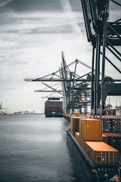 A large cargo ship is docked at an industrial port with towering cranes. Shipping containers are stacked on the ship and along the dock. The sky is overcast, creating a muted ambiance over the water.