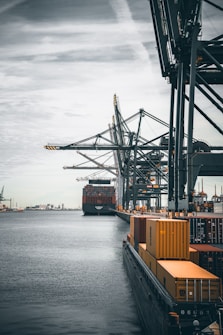 A large cargo ship is docked at an industrial port with towering cranes. Shipping containers are stacked on the ship and along the dock. The sky is overcast, creating a muted ambiance over the water.