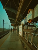 A metro station platform with a high curved roof and metallic supports. Signs in multiple languages are prominently displayed, including the name 'Mayur Vihar Phase-1'. The perspective leads down the length of the platform, where a few commuters walk along the tiled floor. Overhead wires and tracks run parallel to the platform.