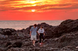 A cinematic capture of a couple walking hand in hand along a charcoal-colored rocky coastline at sunset.