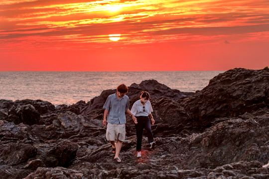 A cinematic capture of a couple walking hand in hand along a charcoal-colored rocky coastline at sunset.