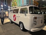 A classic white Volkswagen van is parked on a concrete pathway. It features a red circle with the letters 'AS' on its side. The van has a roof rack loaded with oversized, colorful gift boxes. Nearby, a man in a plaid shirt walks on the left side. Brightly lit shops are visible in the background, along with a decorated Christmas tree.