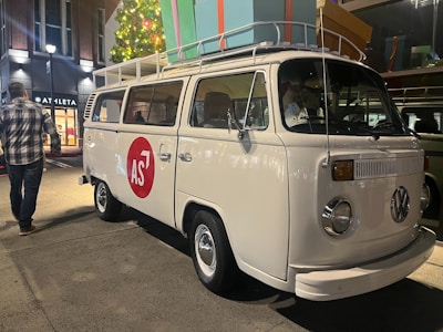 A classic white Volkswagen van is parked on a concrete pathway. It features a red circle with the letters 'AS' on its side. The van has a roof rack loaded with oversized, colorful gift boxes. Nearby, a man in a plaid shirt walks on the left side. Brightly lit shops are visible in the background, along with a decorated Christmas tree.