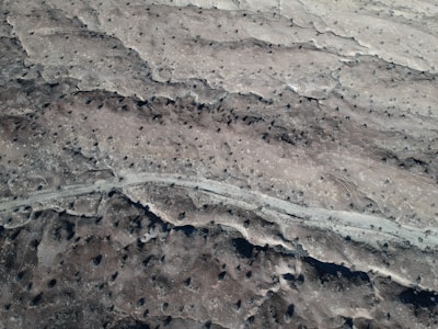 An aerial view reveals a barren, rocky landscape featuring scattered craters and a winding path cutting through the terrain. The area is predominantly grey and brown, with patches of darker and lighter shades giving a textured appearance.