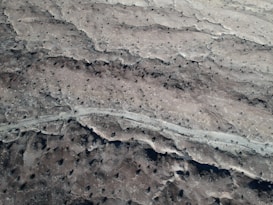 An aerial view reveals a barren, rocky landscape featuring scattered craters and a winding path cutting through the terrain. The area is predominantly grey and brown, with patches of darker and lighter shades giving a textured appearance.