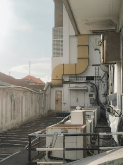 An industrial area with multiple air conditioning units placed along a narrow alley next to a large building. The wall is weathered and shows signs of aging with some wires and pipes directed toward the units. There is a metal railing, and the area appears somewhat neglected. The sky is clear with a light hue, and a distant view of rooftops is visible.