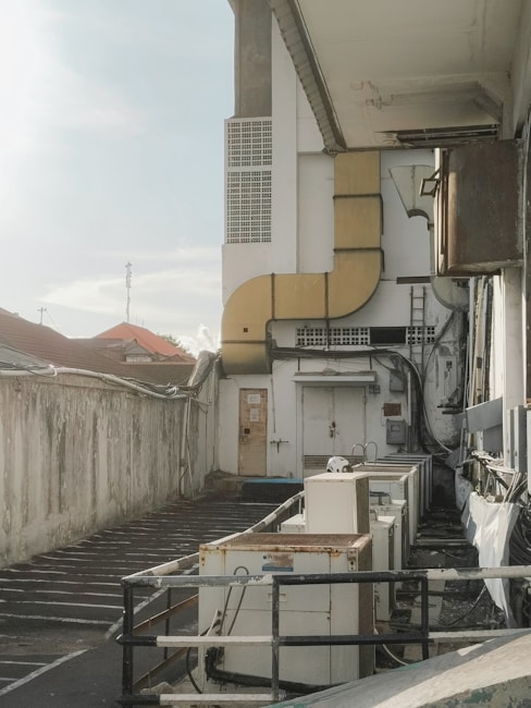 An industrial area with multiple air conditioning units placed along a narrow alley next to a large building. The wall is weathered and shows signs of aging with some wires and pipes directed toward the units. There is a metal railing, and the area appears somewhat neglected. The sky is clear with a light hue, and a distant view of rooftops is visible.