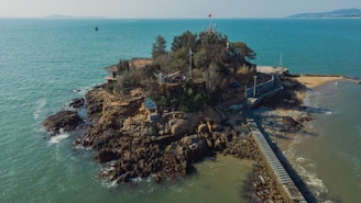 A small rocky island is surrounded by calm turquoise waters. The island features dense vegetation, a few man-made structures including buildings and a bridge that leads to the mainland. There's a lone flagpole with a flag, and the water is gently washing against the rocky shore. The background reveals distant landforms under a clear sky.