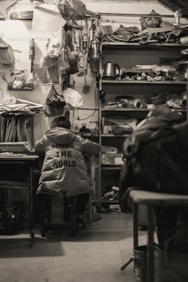 A person wearing a jacket with the words 'Dream the World' is seated in a cluttered room filled with various items. Shelves filled with miscellaneous objects, including a kettle and stacks of materials, are visible. The atmosphere suggests a workshop or storage space.
