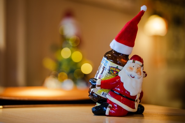 A festive ceramic Santa Claus figurine is holding a brown glass bottle, adorned with a label, on a wooden surface. In the blurred background, colorful circular bokeh lights suggest a Christmas tree setting, adding a warm and cozy holiday atmosphere.