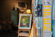An art gallery display featuring a framed painting on an easel in the foreground, depicting an abstract scene with vivid colors. The background includes an antique clock, various artworks, and a warm-lit lamp. The scene is viewed through an open glass door with a yellow 'OPEN' sign.