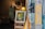 An art gallery display featuring a framed painting on an easel in the foreground, depicting an abstract scene with vivid colors. The background includes an antique clock, various artworks, and a warm-lit lamp. The scene is viewed through an open glass door with a yellow 'OPEN' sign.