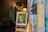 An art gallery display featuring a framed painting on an easel in the foreground, depicting an abstract scene with vivid colors. The background includes an antique clock, various artworks, and a warm-lit lamp. The scene is viewed through an open glass door with a yellow 'OPEN' sign.