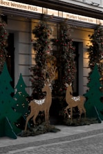 An outdoor holiday display features decorative reindeer figures and Christmas trees adorned with red baubles and lights. The setting includes a building with signage above and cobblestone pavement below, giving it a festive and welcoming atmosphere.