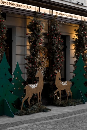 An outdoor holiday display features decorative reindeer figures and Christmas trees adorned with red baubles and lights. The setting includes a building with signage above and cobblestone pavement below, giving it a festive and welcoming atmosphere.
