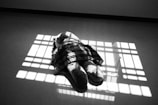 A large stuffed animal lies on the floor within a patterned shadow cast by a window. The stuffed toy is partially covered by a plaid cloth, creating a stark contrast with the floor.