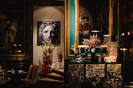 A stylish and elegant interior featuring a piece of art depicting a classical-style face. The room includes a rich color palette with teal and gold accents. Decorative items such as a vase with pampas grass and a poinsettia plant are placed below the artwork. A large mirror reflects part of the room, adding depth. A dresser is adorned with festive decorations, including a miniature tree and red berries.