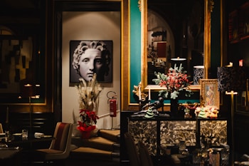 A stylish and elegant interior featuring a piece of art depicting a classical-style face. The room includes a rich color palette with teal and gold accents. Decorative items such as a vase with pampas grass and a poinsettia plant are placed below the artwork. A large mirror reflects part of the room, adding depth. A dresser is adorned with festive decorations, including a miniature tree and red berries.