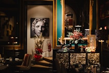 A stylish and elegant interior featuring a piece of art depicting a classical-style face. The room includes a rich color palette with teal and gold accents. Decorative items such as a vase with pampas grass and a poinsettia plant are placed below the artwork. A large mirror reflects part of the room, adding depth. A dresser is adorned with festive decorations, including a miniature tree and red berries.