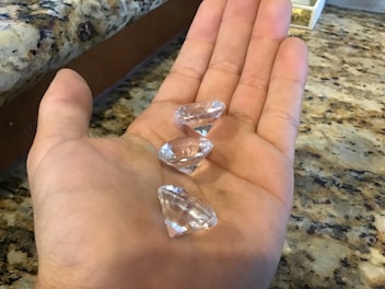 A hand holds three transparent, faceted stones resembling diamonds. The background features a granite countertop with a speckled pattern.