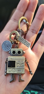 A metallic robot keychain is held in the palm of a hand. The robot features a retro design with large circular eyes, a square body covered in various patterns, and movable limbs. Attached to the keychain is a keyring and a small metal tag engraved with text, accompanied by a circular metal piece with a spiral pattern.