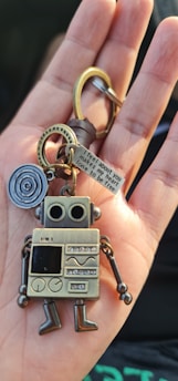 A metallic robot keychain is held in the palm of a hand. The robot features a retro design with large circular eyes, a square body covered in various patterns, and movable limbs. Attached to the keychain is a keyring and a small metal tag engraved with text, accompanied by a circular metal piece with a spiral pattern.