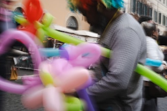 A person with a colorful wig is holding balloons of various shapes and colors, including pink, green, purple, and red. The scene appears lively and dynamic, with a slightly blurred effect indicating motion. In the background, there are other people and buildings, suggesting a busy street or marketplace environment.