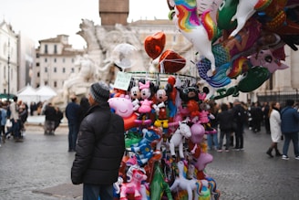A street vendor stands beside a colorful display of various balloons in a public square. The assortment features cartoon characters and animals like Mickey Mouse, Peppa Pig, and unicorns. In the background, a crowd of people walks around, with historic buildings and a fountain visible.