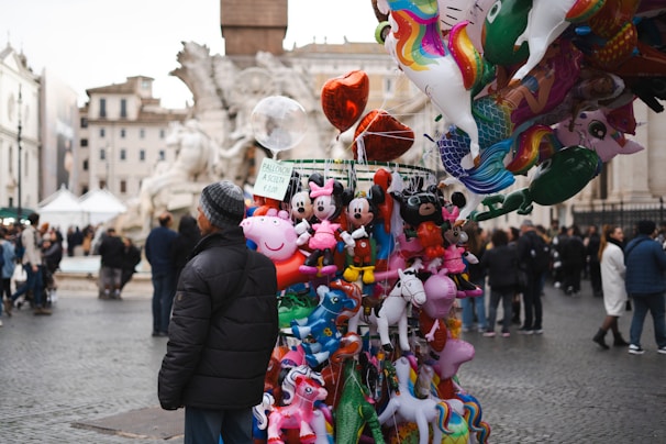 A street vendor stands beside a colorful display of various balloons in a public square. The assortment features cartoon characters and animals like Mickey Mouse, Peppa Pig, and unicorns. In the background, a crowd of people walks around, with historic buildings and a fountain visible.