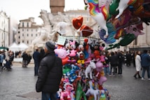 A street vendor stands beside a colorful display of various balloons in a public square. The assortment features cartoon characters and animals like Mickey Mouse, Peppa Pig, and unicorns. In the background, a crowd of people walks around, with historic buildings and a fountain visible.