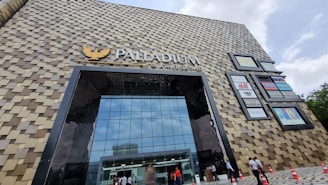 The image displays a large, modern building with a unique facade composed of a pattern of square tiles. The building has a prominent glass entrance, and several brand logos are visible on the side. People are walking in and out, and traffic cones are lined up near the entrance.