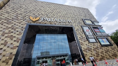 The image displays a large, modern building with a unique facade composed of a pattern of square tiles. The building has a prominent glass entrance, and several brand logos are visible on the side. People are walking in and out, and traffic cones are lined up near the entrance.