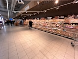 A supermarket aisle with shelves stocked with various packaged goods. Two people are standing in the middle of the aisle, and several shopping carts are visible. The shelves along the wall display a variety of items including snacks and packaged foods. Overhead signs and lights are present, and the flooring is made of large tiles.
