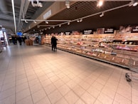 A supermarket aisle with shelves stocked with various packaged goods. Two people are standing in the middle of the aisle, and several shopping carts are visible. The shelves along the wall display a variety of items including snacks and packaged foods. Overhead signs and lights are present, and the flooring is made of large tiles.