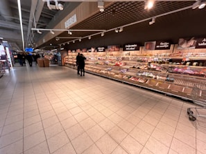 A supermarket aisle with shelves stocked with various packaged goods. Two people are standing in the middle of the aisle, and several shopping carts are visible. The shelves along the wall display a variety of items including snacks and packaged foods. Overhead signs and lights are present, and the flooring is made of large tiles.
