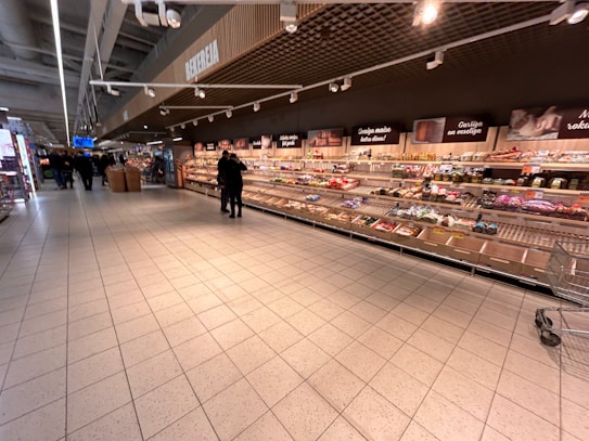 A supermarket aisle with shelves stocked with various packaged goods. Two people are standing in the middle of the aisle, and several shopping carts are visible. The shelves along the wall display a variety of items including snacks and packaged foods. Overhead signs and lights are present, and the flooring is made of large tiles.
