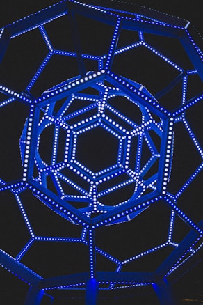 A geometric structure of interconnected lines forms a complex polyhedral shape, illuminated in vibrant blue lights against a dark background. The intricate pattern and the symmetry of the structure are visually striking.