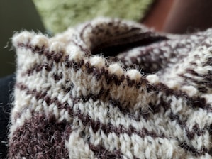 A close-up view of a knitted fabric featuring a pattern of alternating brown and beige stripes. The texture appears soft and fuzzy, with visible individual yarn strands and a warm, cozy appearance. The background is blurred, suggesting a shallow depth of field.