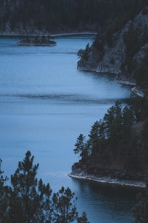 A serene lake bordered by rocky cliffs and dense pine trees, creating a tranquil and secluded atmosphere. The water is a deep blue, reflecting the surrounding nature and blending seamlessly with the dark hues of the trees and rocks.